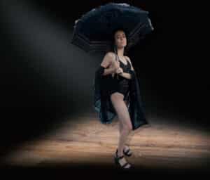 Model wearing black with black umbrella under a spotlight in a dark room.